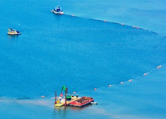 Cap material consisting of larger rocks is applied by backhoe from a barge (foreground). The cap creates a new habitat layer on the lake bottom.