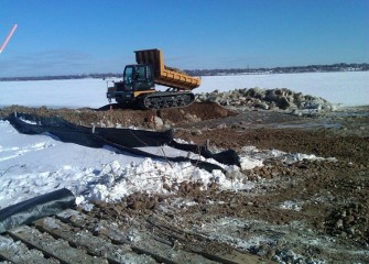 A rubber-tracked dump truck suitable for moving over uneven terrain delivers clean soil to the edge of the lake.