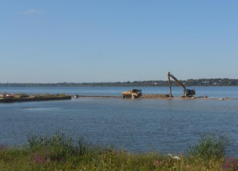 Near the mouth of Harbor Brook, workers reach shallow areas over heavy equipment mats used to create temporary access roads. A Great Egret can be seen at the shore’s edge.